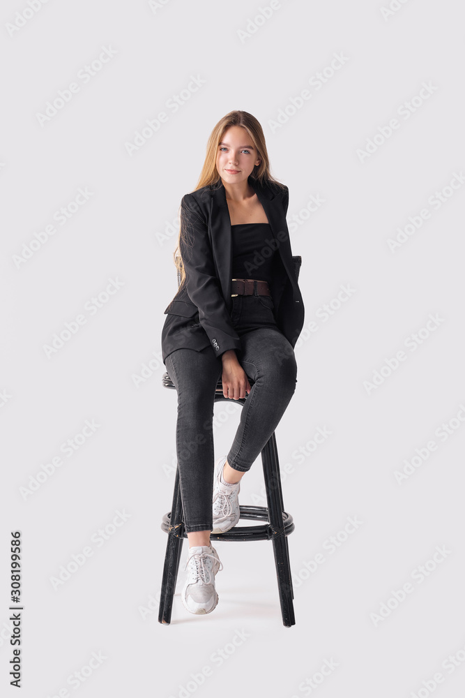 Fototapeta premium Full length portrait of beautiful young girl sitting on tall chair in black jeans and jacket over gray background