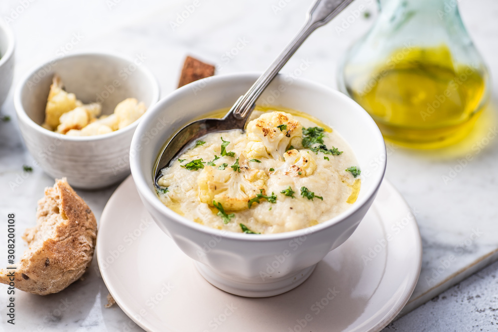 Roasted cauliflower cream soup on white kitchen table, served with fresh parsley, olive oil and bread. Roasted cauliflower cream soup on white kitchen table, served with fresh parsley, olive oil and bread.