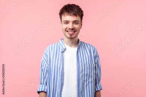 Portrait of positive young brown-haired man with small beard and mustache in casual striped shirt, looking at camera with toothy smile, optimistic view. indoor studio shot isolated on pink background