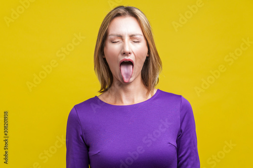 Portrait of positive carefree woman with fair hair in purple dress standing with closed eyes and demonstrating tongue, naughty disobedient expression. indoor studio shot isolated on yellow background