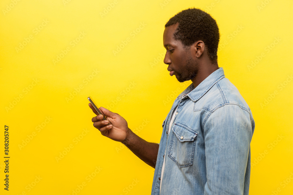 Side view of serious man in denim shirt using cellphone, typing text message or dialing number with calm attentive expression, technology concept. indoor studio shot isolated on yellow background