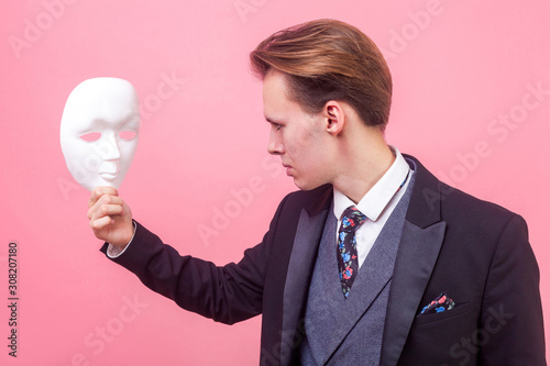 Portrait of upset serious young businessman in elegant suit and with stylish haircut looking attentively at white mask with gloomy depressed expression. indoor studio shot isolated on pink background