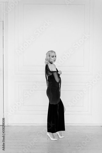 slim girl with blond hair is standing in a long evening dress on a white background. Luxury, rich lifestyle. Fashion shot. Interior in classical style. Black and white photo