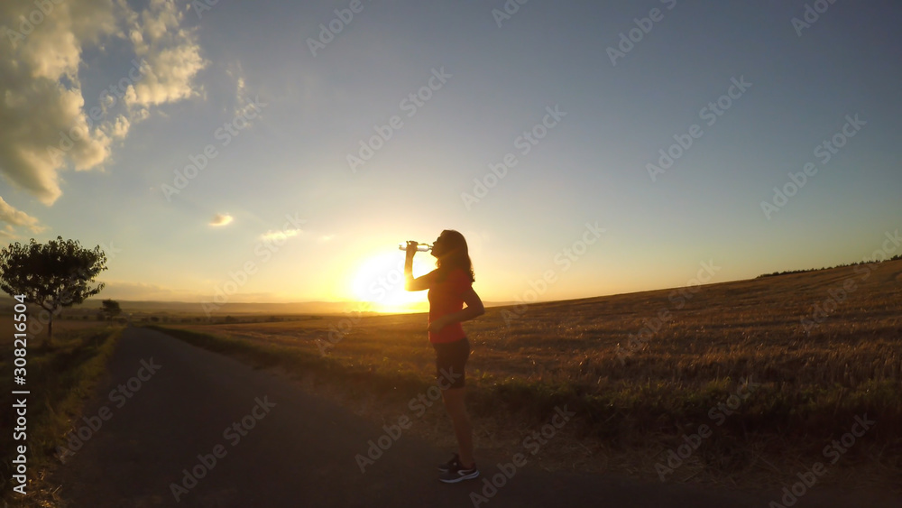 Running at sunset. A young woman drinking water during a running exercise. (Intentionally uneven horizon line - fish eye effect)