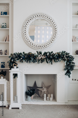 View over decorated artificial fireplace with decorations inside. Handmade paper stars, candles inside. Beautiful coniferous wreath on the top of fireplace. Decorative round mirror over the fireplace.