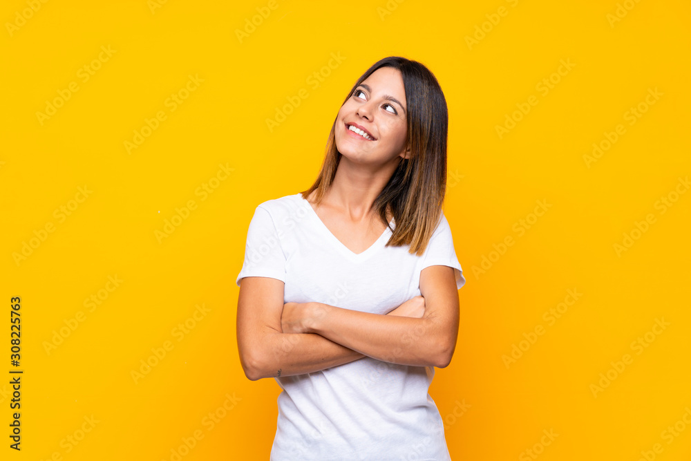 Young woman over isolated yellow background looking up while smiling - obrazy, fototapety, plakaty 