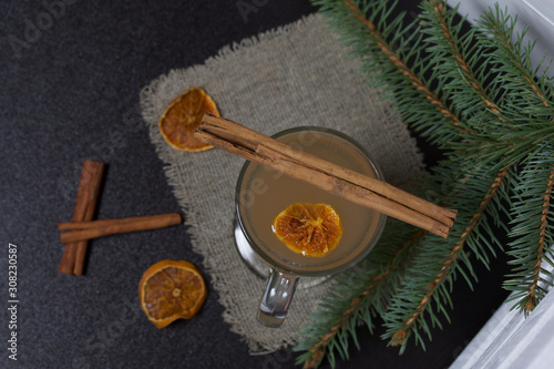 A glass of orange drink stands on a piece of linen. A slice of dried orange floats in it. Nearby are cinnamon sticks and dried oranges and a spruce branch.