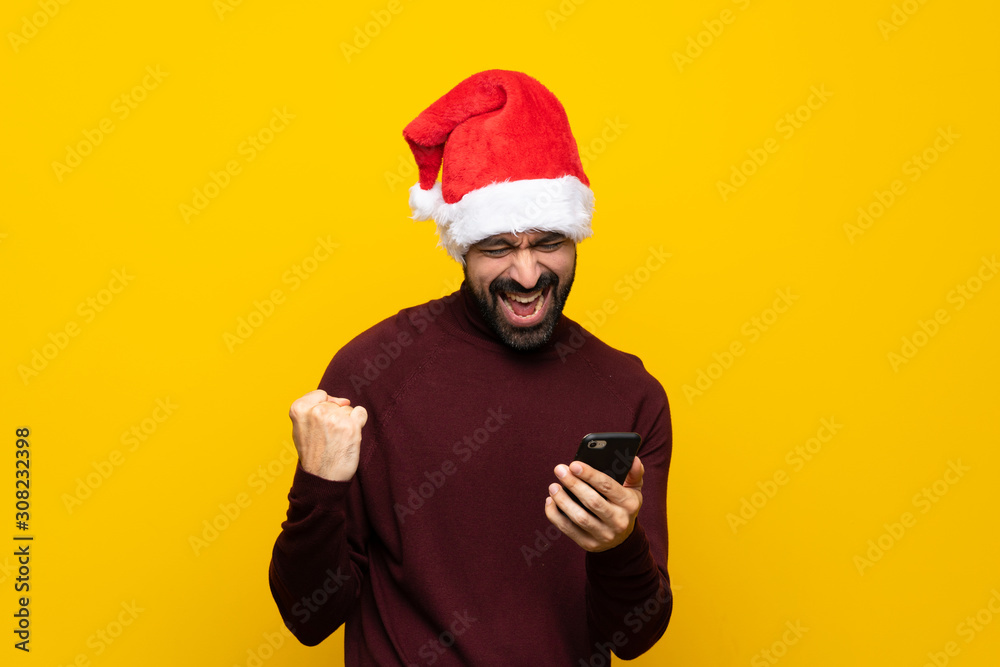 Man with christmas hat over isolated yellow background with phone in victory position Man with christmas hat over isolated yellow background with phone in victory position