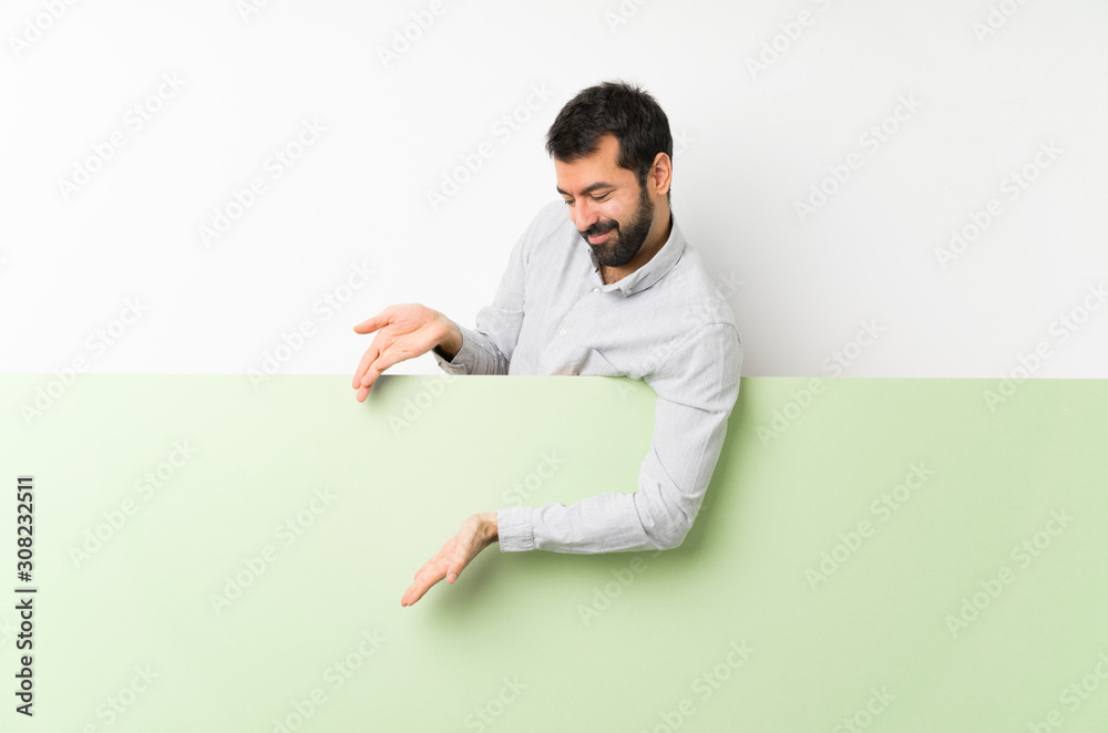 Young handsome man with beard holding a big green empty placard extending hands to the side for inviting to come Young handsome man with beard holding a big green empty placard extending hands to the side for inviting to come