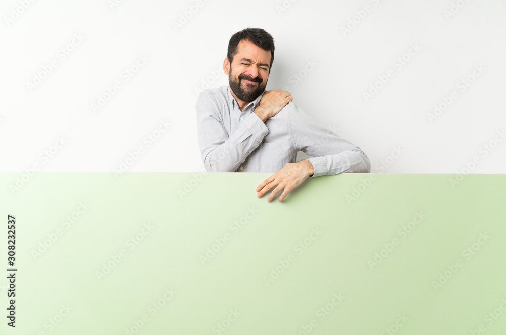 Young handsome man with beard holding a big green empty placard suffering from pain in shoulder for having made an effort Young handsome man with beard holding a big green empty placard suffering from pain in shoulder for having made an effort