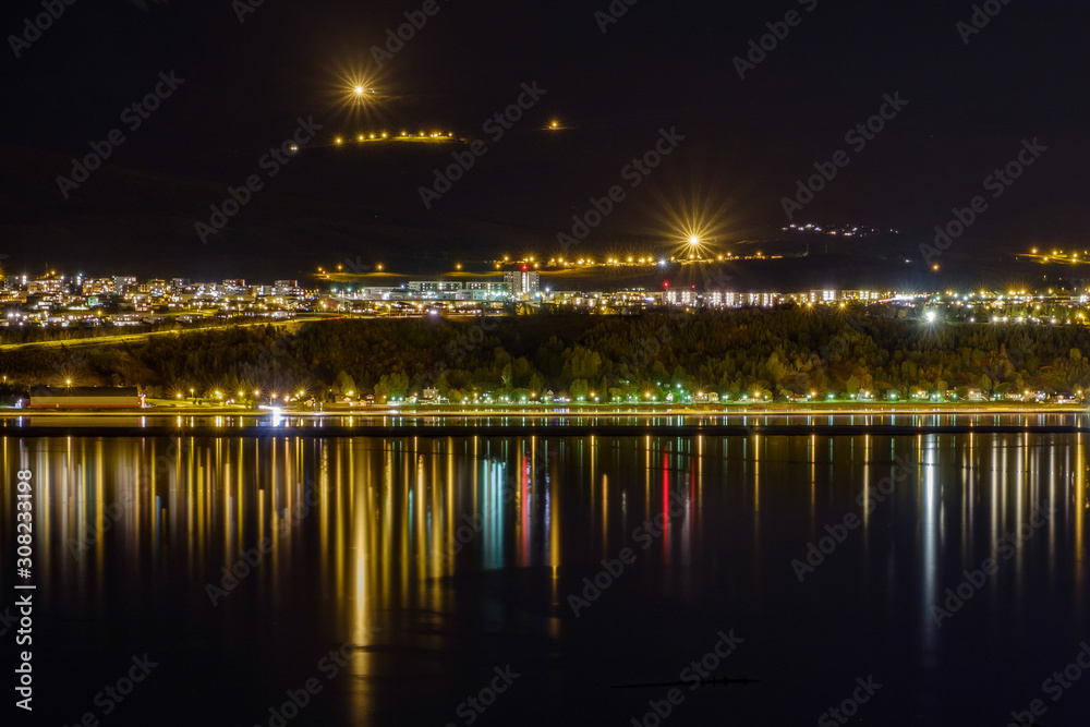Akureyri at night, in the foreground mirroring water with the lights of the city reflecting in the water,