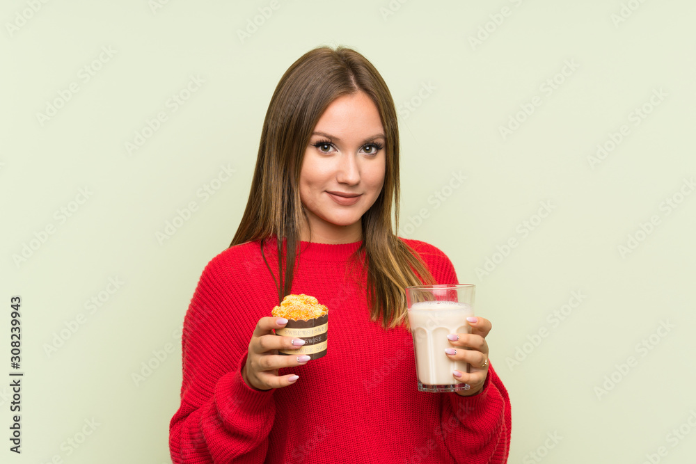 Teenager girl holding a glass of milk and a muffin over isolated green background Teenager girl holding a glass of milk and a muffin over isolated green background