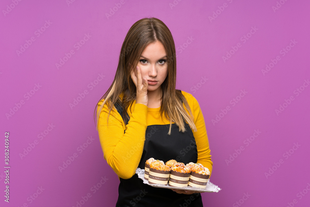 Teenager girl holding lots of different mini cakes over isolated purple background unhappy and frustrated