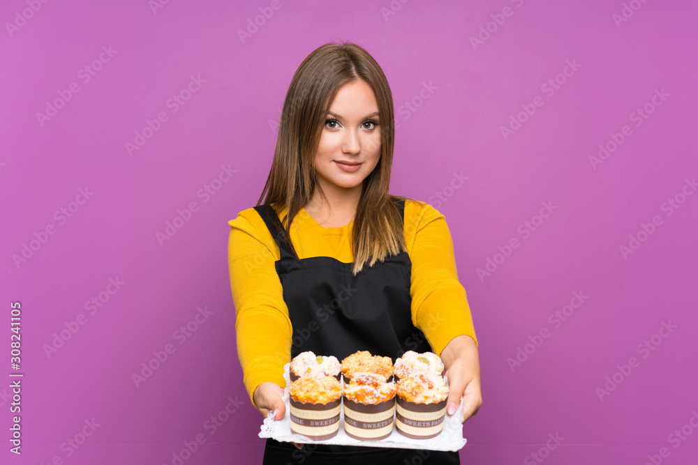 Teenager girl holding lots of different mini cakes over isolated purple background Teenager girl holding lots of different mini cakes over isolated purple background