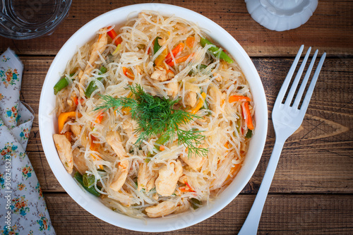 Funchosa with vegetables and chicken on a white plate on a wooden table, selective focus