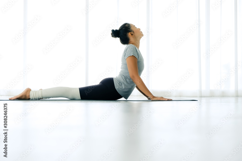 Obraz premium Asian yogi woman practicing yoga concept, stretching in Cobra exercise, Bhujangasana pose, working out, indoor full length, white yoga studio ,concept of healthy life and natural balance