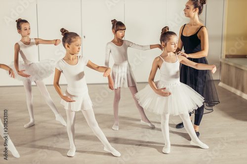 young caucasian ballerina in black suit teaching little children in white suits, practicing and coaching them in studio, correct body position