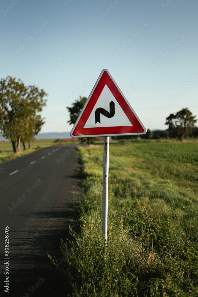 Photo of illuminated and warning road sign - double curve next to the road on meadow. Double Bend warning road sign on country road with trees and beautiful landscape on background - European sign.