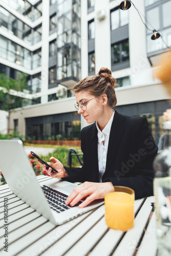 Young business woman wearing glasses at cafe using laptop and drinking coffee. Businesswoman drinking coffee at cafe and hold smartphone