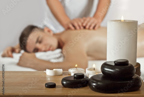 A naked man covered in a towel is waiting for a massage. The guy lies on the table on a white background. Massage stones and candles in the foreground.