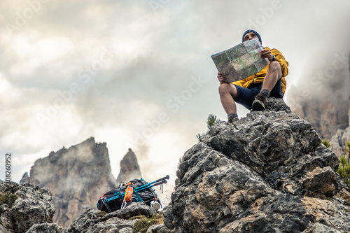 Young man hiker sitting on stone mountain reading map, with cloudy sky and fog. Yellow jacket, backpack, black beard and beanie. Traveling dolomites, Italy.