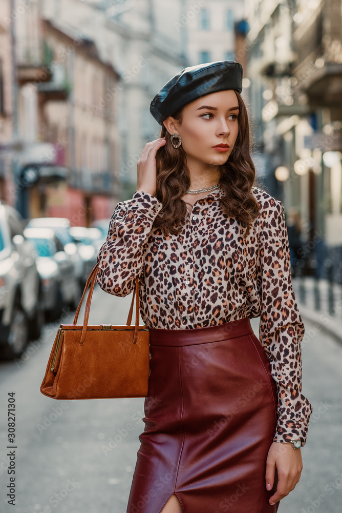Outdoor fashion portrait of young elegant model, woman wearing black beret,  leopard print blouse, faux leather dark red skirt, holding stylish brown  bag, handbag, walking in street of European city Photos
