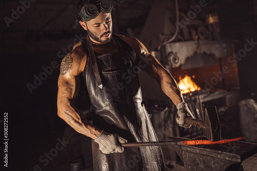 young bearded man in leather uniform heats the metal on fire isolated in workshop