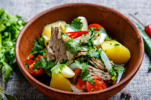 Hashlama in a clay plate. Ingredients: Beef ribs, potatoes, herbs, garlic. The plate stands on a textured gray canvas. Around lie pepper red and green pods, pepper peas, tomatoes, Bay leaf, parsley photo