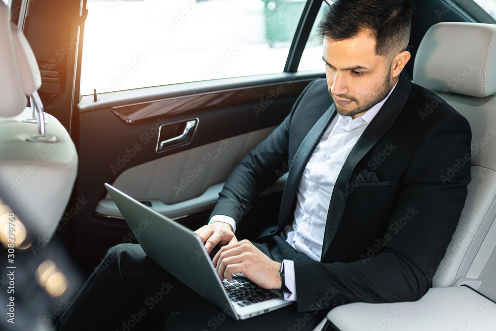 handsome confident businessman typing on laptop, busy young caucasian man sit in luxurious car wearing tuxedo