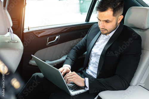 handsome confident businessman typing on laptop, busy young caucasian man sit in luxurious car wearing tuxedo © alfa27