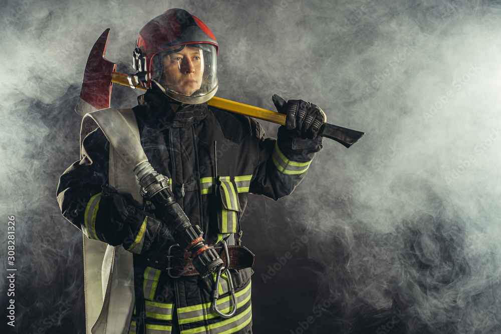 Naklejka premium young caucasian fireman holding hammer, risking his life to save people from fire, wearing protective uniform