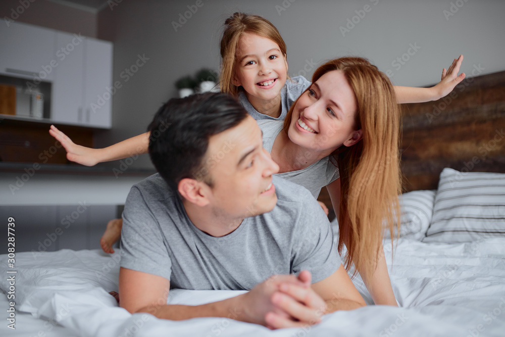 lovely caucasian parents lying on bed with their kid girl, wearing casual clothes, happy smiling family together at home