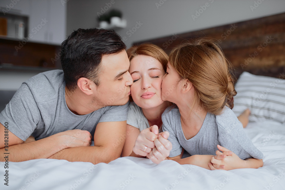 portrait of beautiful happy family lying on bed together, mother father and kid girl kissing hugging and laughing. indoors