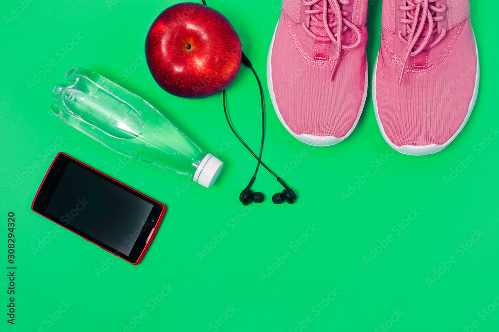 Pink sport shoes, earphones, red apple, smartphone and bottle of water on a green background. Concept healthy lifestyle, sport and diet. 