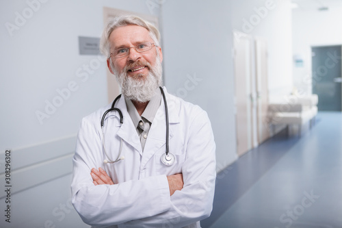 Happy senior male doctor smiling to the camera, standing in hallway of his clinic, copy space