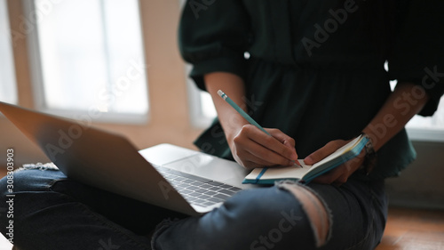 Closeup woman writing notepad and put laptop on leg with sitting on the floor.