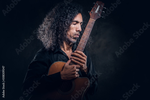 In dark photo studio curly middle aged man is posing with guitar for photographer.