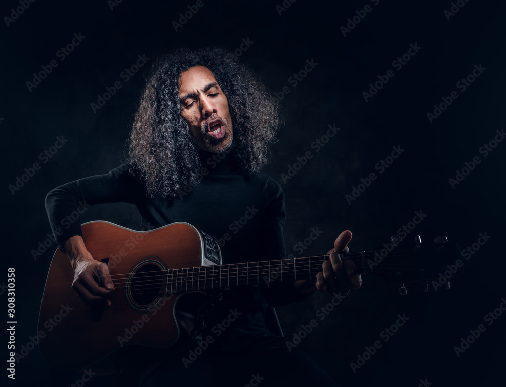 Handsome man with long hair is playing acoustic guitar at dark studio.