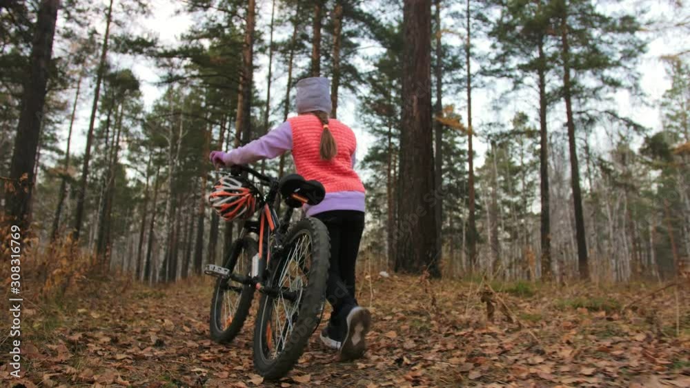 One caucasian children walk with bike in autumn park. Little girl walking black orange cycle in forest. Kid goes do bicycle sports. Biker motion ride with backpack and helmet. Mountain bike hardtail. One caucasian children walk with bike in autumn park. Little girl walking black orange cycle in forest. Kid goes do bicycle sports. Biker motion ride with backpack and helmet. Mountain bike hardtail.