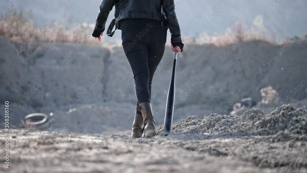 view from the back of a brutal woman walking along a wasteland with a baseball bat