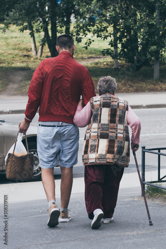 Young man helps old lady with a stick to carry her bag and cross the road