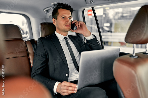 Young handsome businessman is sitting in luxury car. Serious handsome man in suit is working with laptop and talking on smart phone while being in trip. © Valerii Apetroaiei