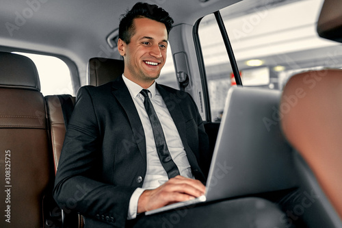 Young handsome businessman is sitting in luxury car. Man in suit is working with laptop while being in trip. © Valerii Apetroaiei