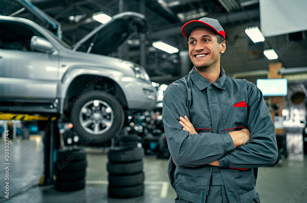 Handsome auto service mechanic in uniform is standing on the background of car with open hood, smiling and looking away. Car repair and maintenance. - obrazy, fototapety, plakaty 