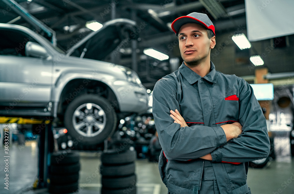 Handsome auto service mechanic in uniform is standing on the background of car with open hood and looking away. Car repair and maintenance. Handsome auto service mechanic in uniform is standing on the background of car with open hood and looking away. Car repair and maintenance.