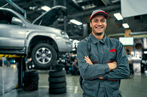 Obraz na płótnie Handsome auto service mechanic in uniform is standing on the background of car with open hood, smiling and looking at camera