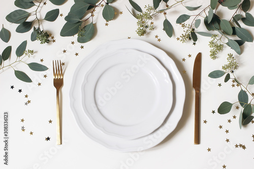 Christmas, New Year table place setting. Golden cutlery, porcelain plate, eucalyptus branches and golden confetti stars isolated on white background. Winter holidays background. Flat lay, top view.