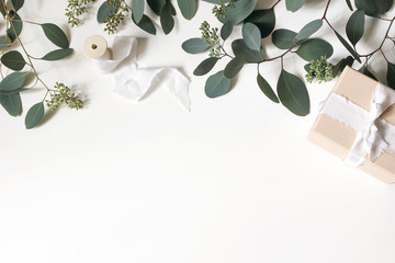 Creative composition made of green berry Eucalyptus populues leaves and branches, gift box and wooden spool with silk ribbon isolated on white background. Wedding styled photo, flat lay, top view.