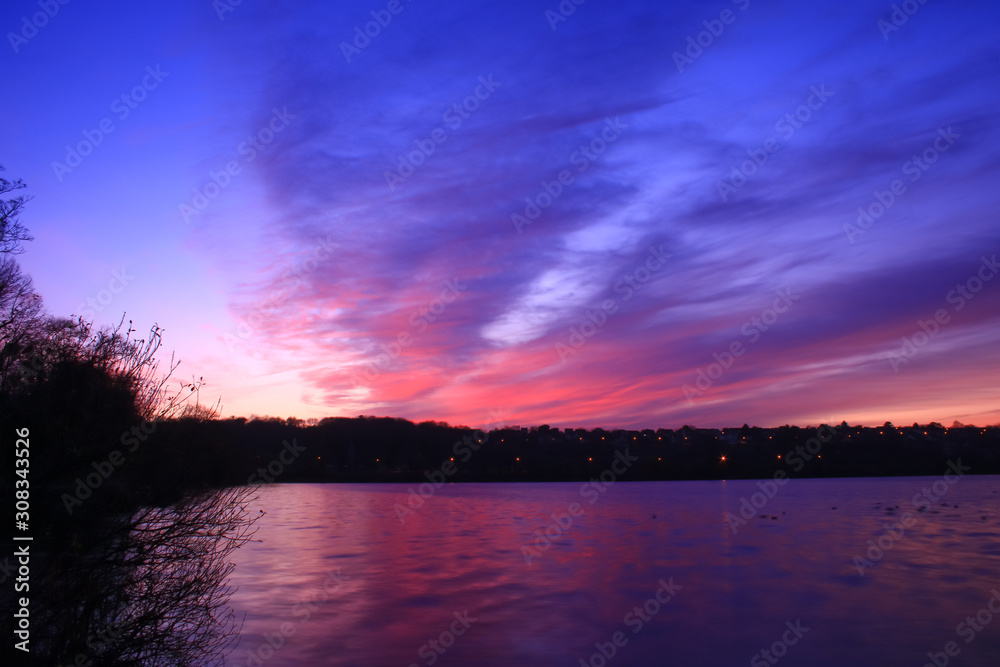 Colorful sunset on the lake. Pink and blue cumulonimbus clouds that are reflected in the water. Silhouette of vegetations in foreground. Light of the city in the background.