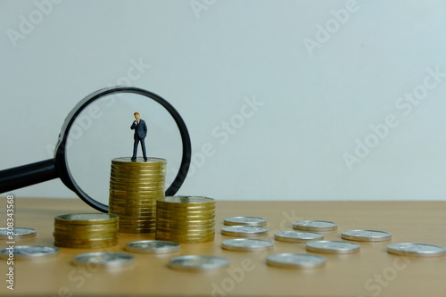 Miniature people business concept - a businessman standing on coin stack with magnifier, searching for financial solution / strategy